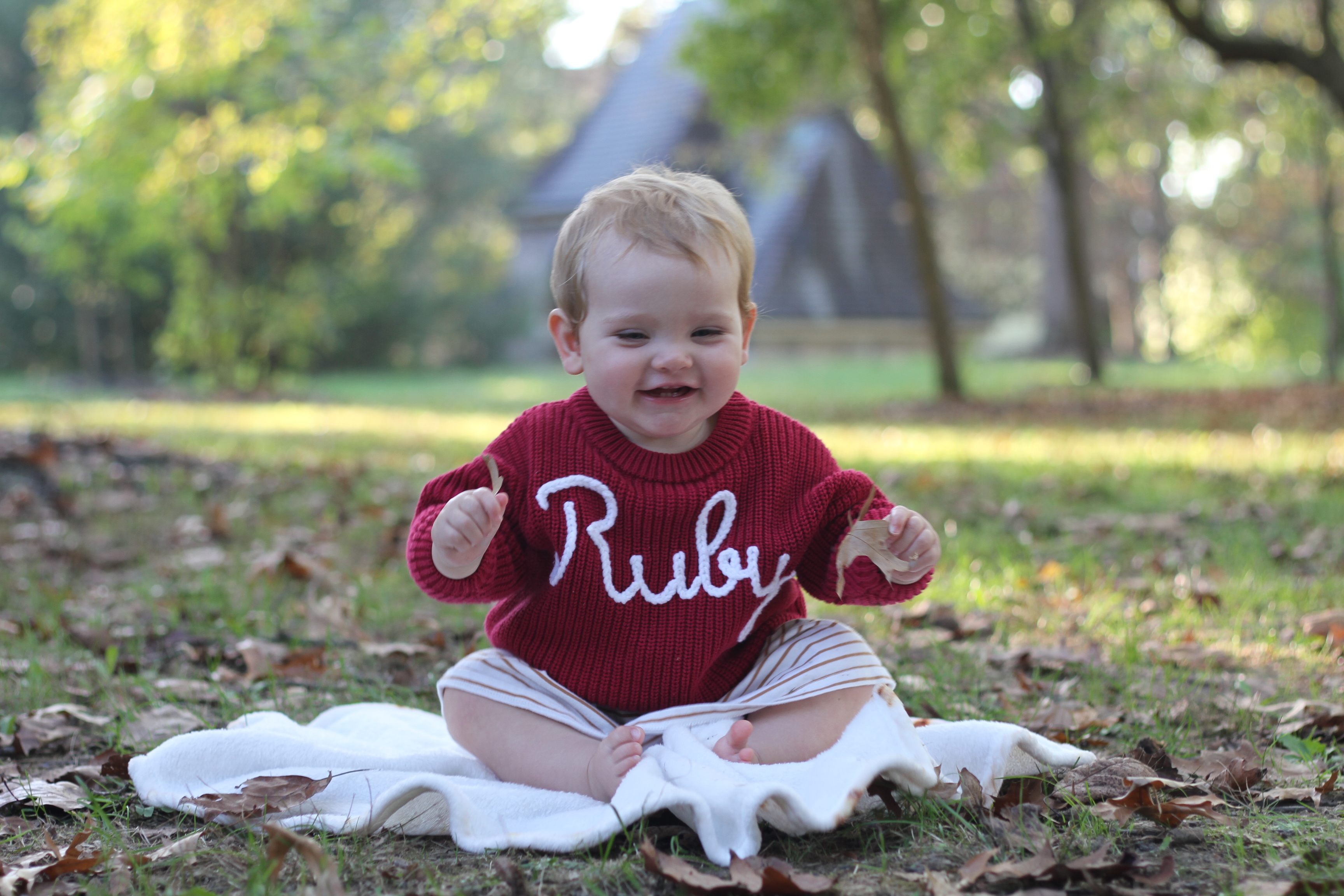 Smiling toddler in a red Ruby sweater sitting on a blanket among autumn leaves